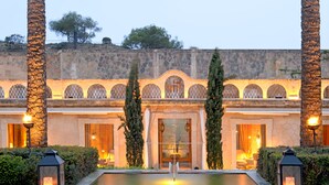 A courtyard with a pool, palm trees, and a stone building with arched windows and a fountain. - Cap Rocat (Llucmajor)