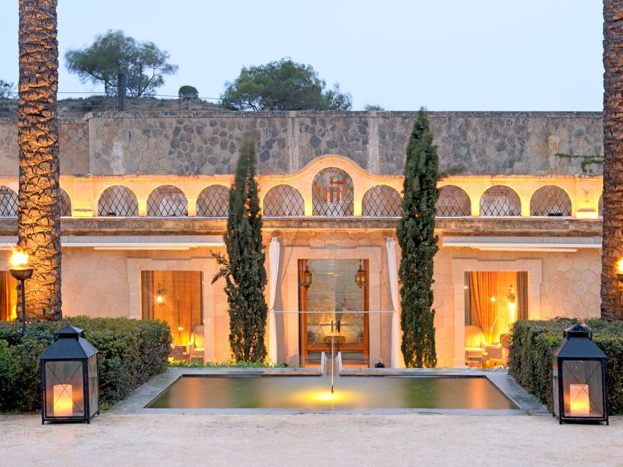 A courtyard with a pool, palm trees, and a stone building with arched windows and a fountain.