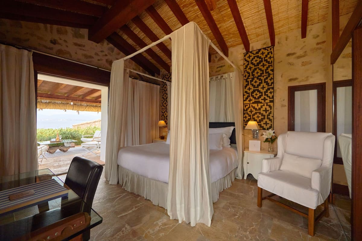 A bedroom with a four-poster bed, a white armchair, a small table with a lamp, and a view of the outdoors.