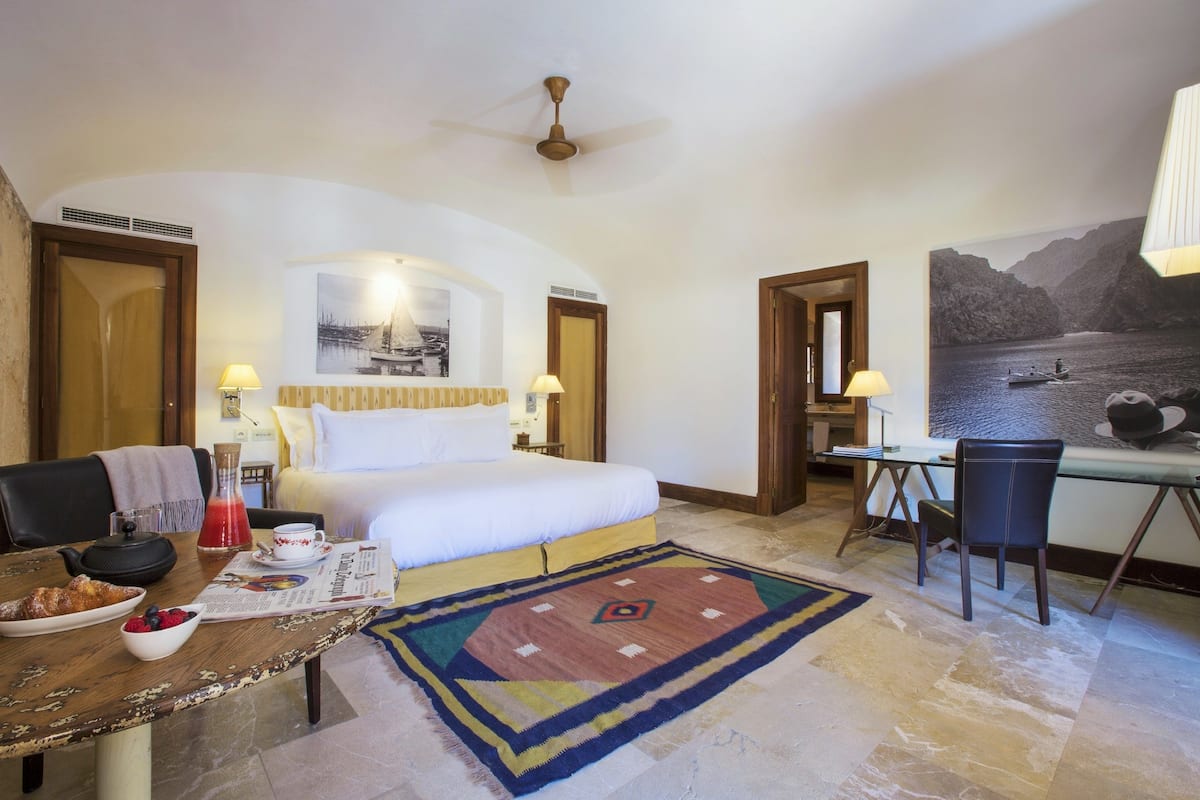 A hotel room with a bed, desk, and dining table. There is a painting on the wall and a ceiling fan.