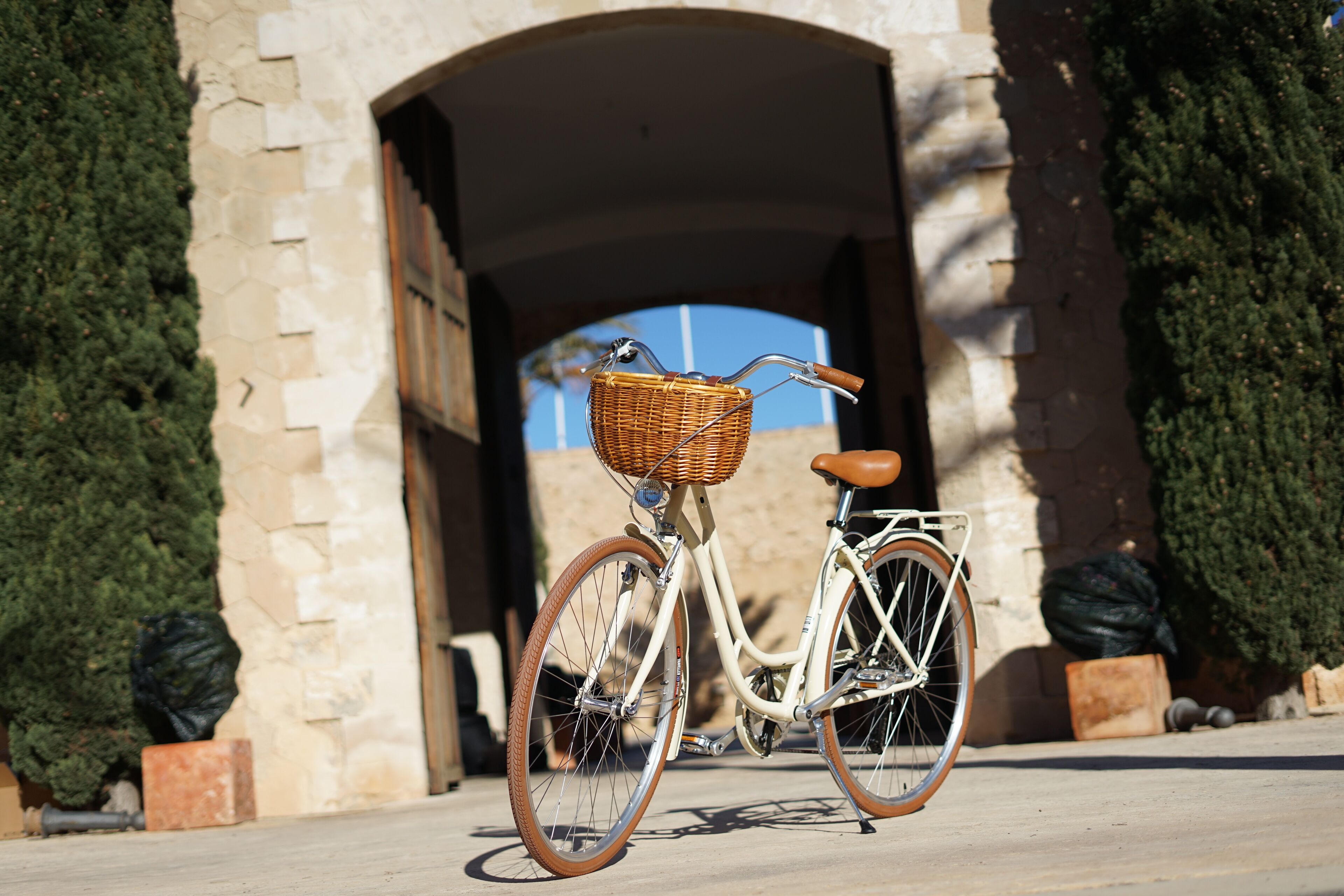 A bicycle with a wicker basket and brown seat parked in front of a stone archway.
