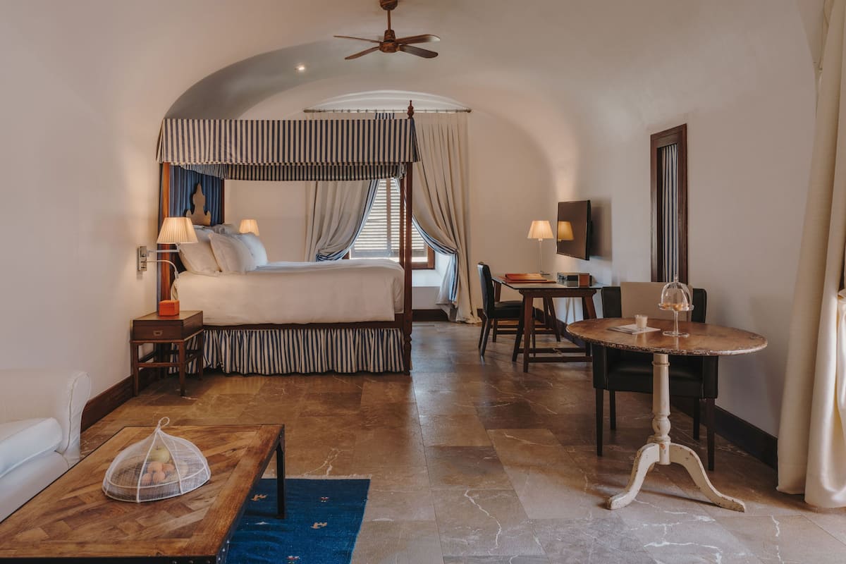 A hotel room with a four-poster bed, a desk with a chair, a dining table, and a ceiling fan.