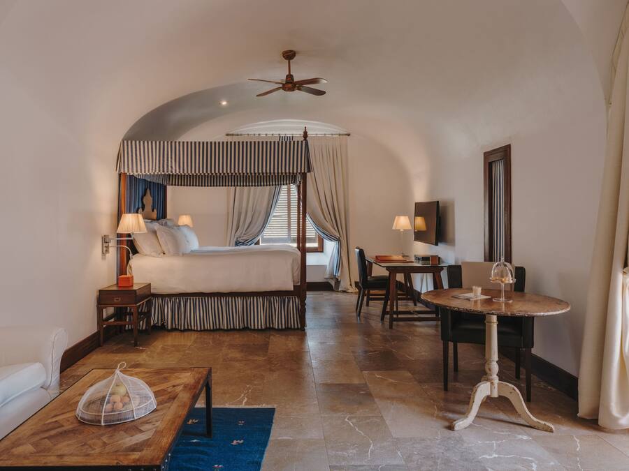 A hotel room with a four-poster bed, a desk with a chair, a dining table, and a ceiling fan.