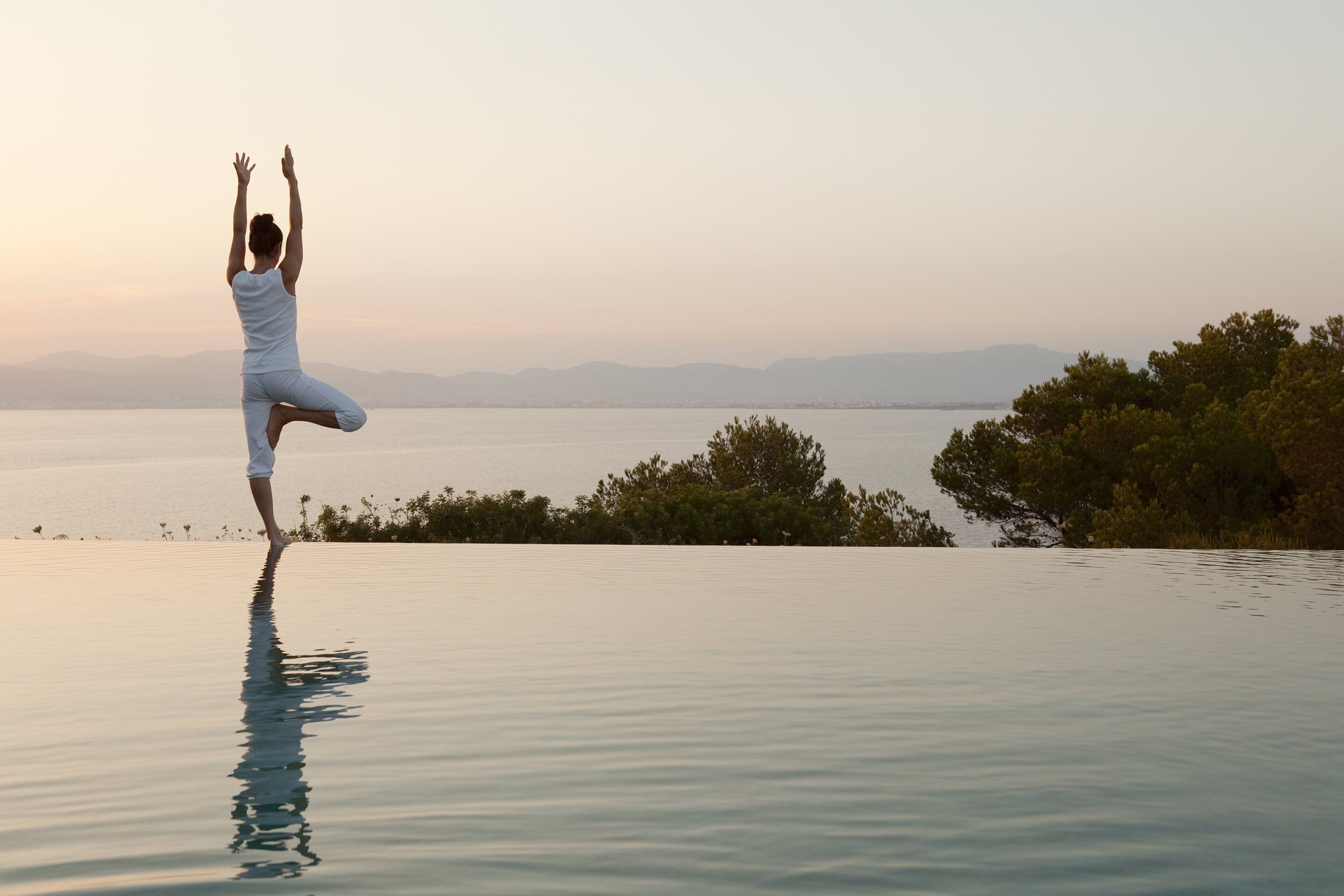 Una persona realizando una postura de yoga junto a una piscina, con vistas a un cuerpo de agua tranquilo y montañas lejanas.