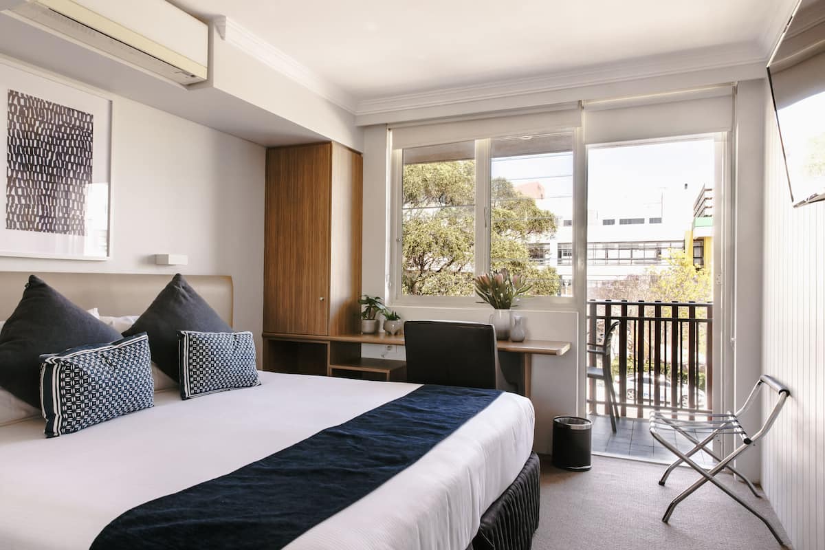 Ein Hotelzimmer mit Bett, Schreibtisch, Stuhl, einem Fenster mit Blick auf Bäume und Gebäude sowie einem Balkon mit Sitzgelegenheit.