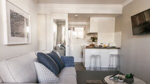 Una sala moderna con un sofá gris, taburetes transparentes, una mesa redonda de centro y una zona de cocina con armarios blancos y un televisor montado en la pared.