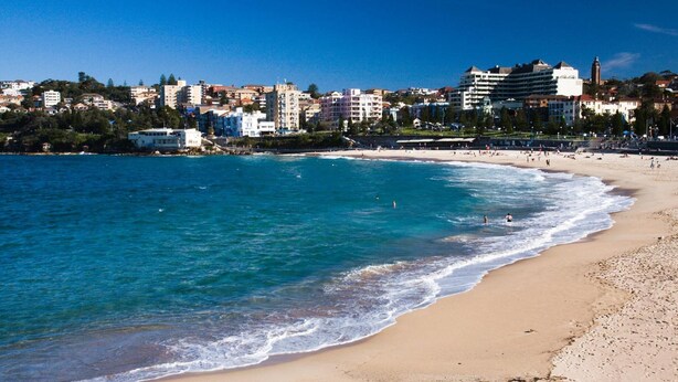 Una playa con agua azul cristalina, una orilla arenosa y un paisaje urbano al fondo.