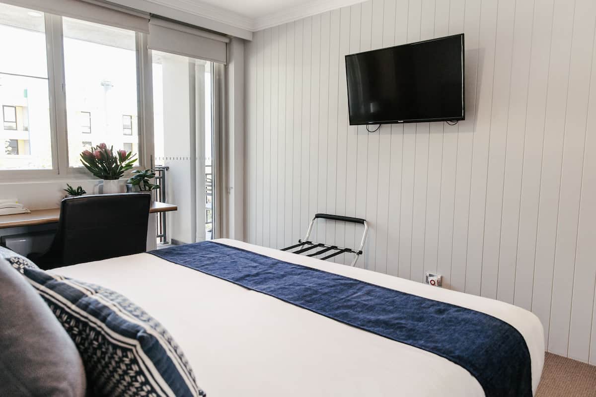 Ein Hotelzimmer mit einem Bett, einem Schreibtisch mit Stuhl, einem an der Wand befestigten Fernseher und einem Fenster mit Blick auf Gebäude.