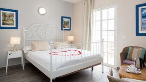 Un dormitorio con una cama de metal blanco, una mesita de noche con lámpara y una silla de mimbre con bandeja.