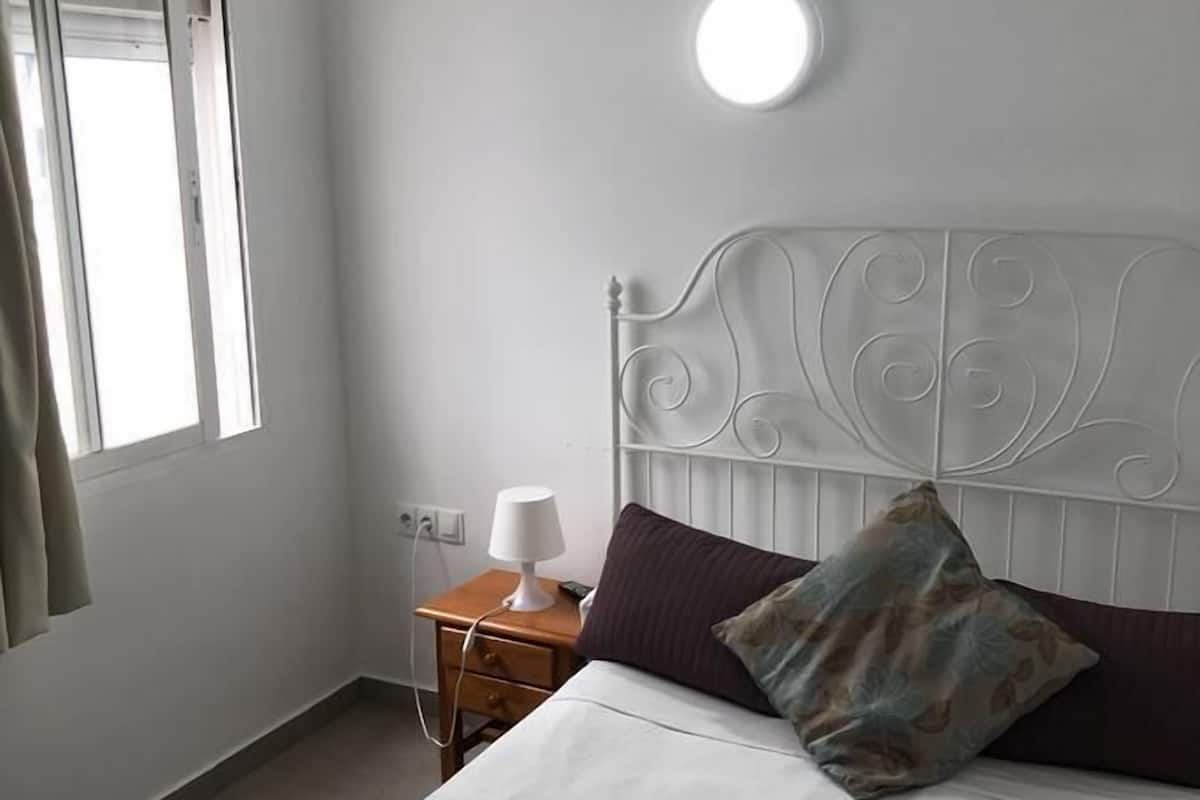 A bedroom with a bed, a nightstand with a lamp, a window, and a wall-mounted light.
