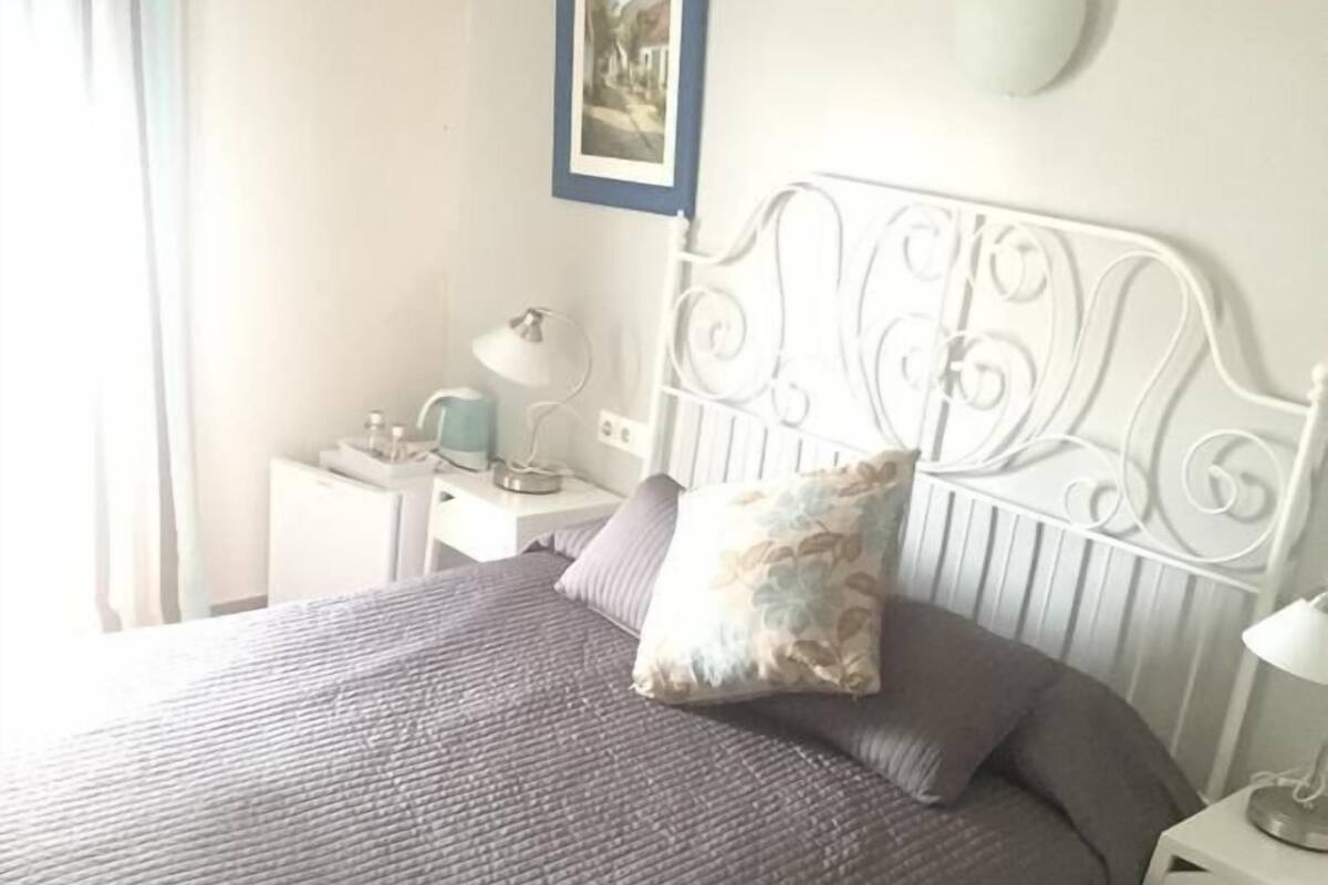 A neatly made bed with a grey quilt and a decorative pillow, a white metal headboard, a bedside table with a lamp, and a framed picture on the wall.