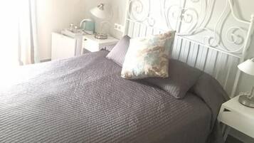 A neatly made bed with a grey quilt and a decorative pillow, a white metal headboard, a bedside table with a lamp, and a framed picture on the wall.