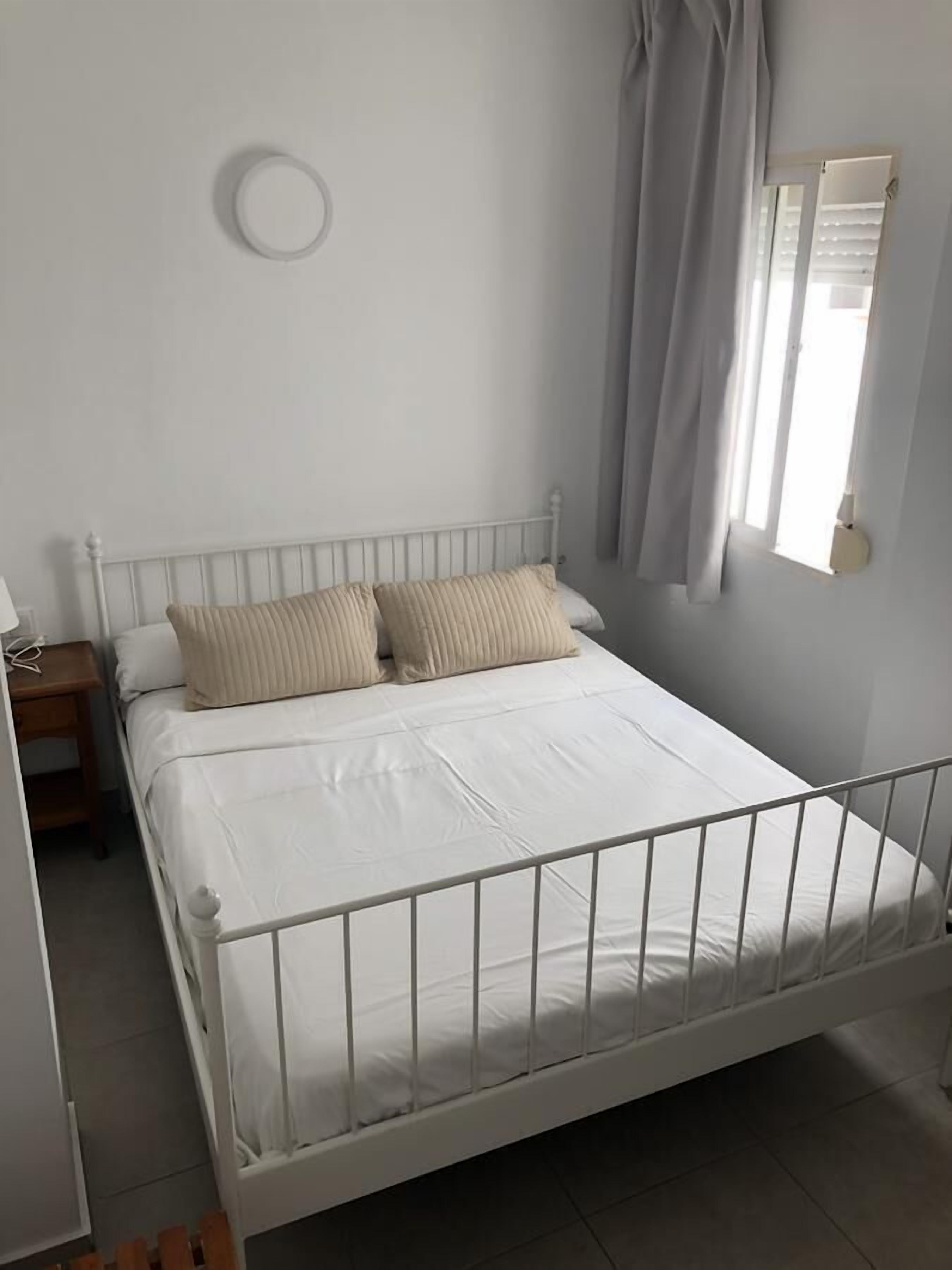 Una cama individual con estructura metálica blanca, dos almohadas color beige y un colchón blanco. Hay una mesita de noche con una lámpara, y una ventana con cortinas.