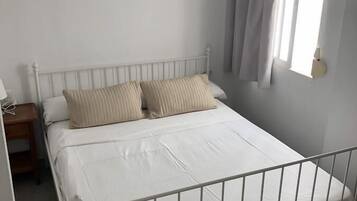 Un llit individual amb estructura metàl·lica blanca, dos coixins beix i matalàs blanc. Hi ha una petita taula de nit amb una làmpada, i una finestra amb cortines.