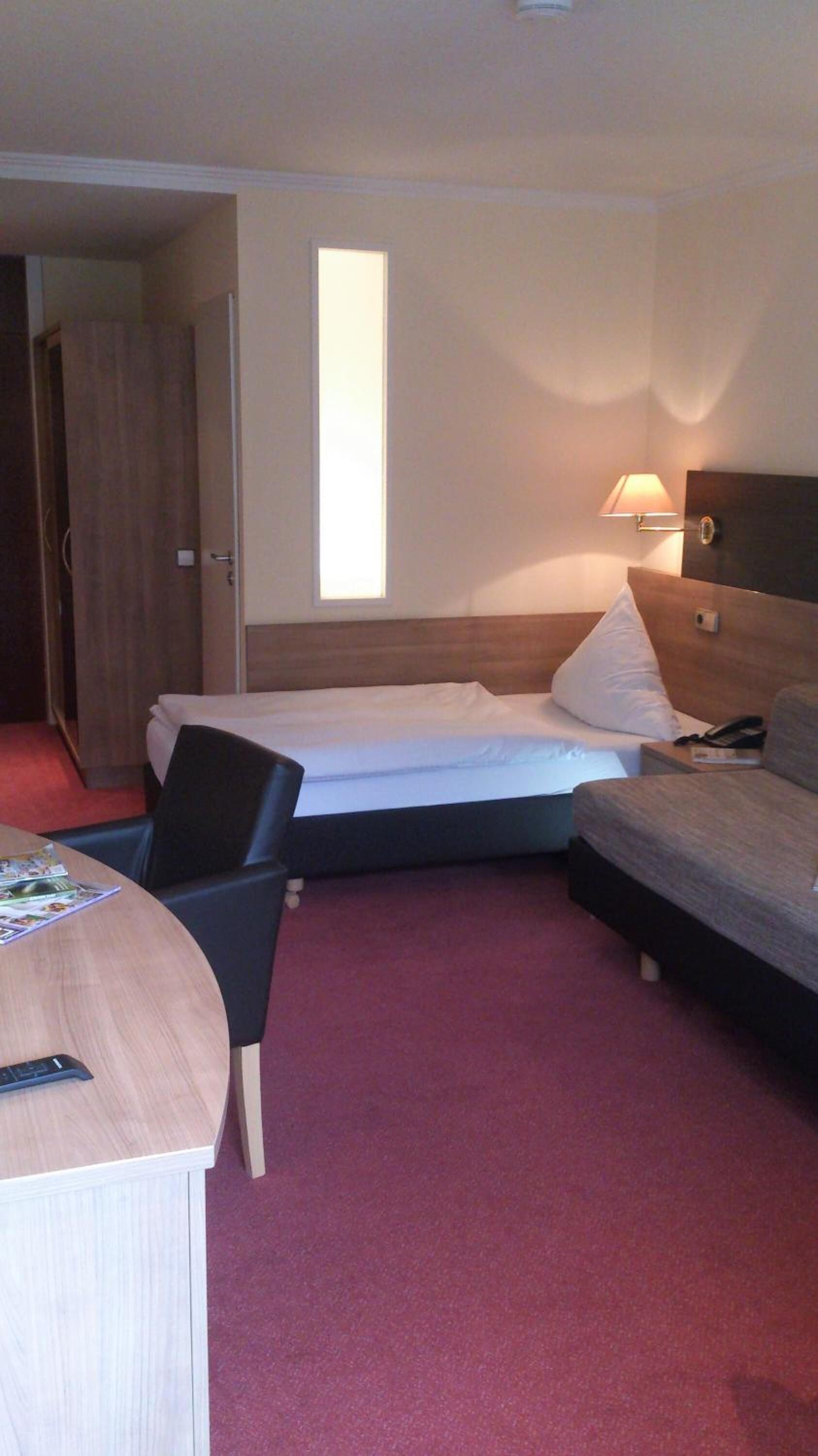 A hotel room with a bed, a desk with a chair, a bedside table, and a lamp.