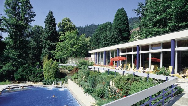 Una piscina con gente, rodeada de vegetación exuberante y un edificio con terraza.