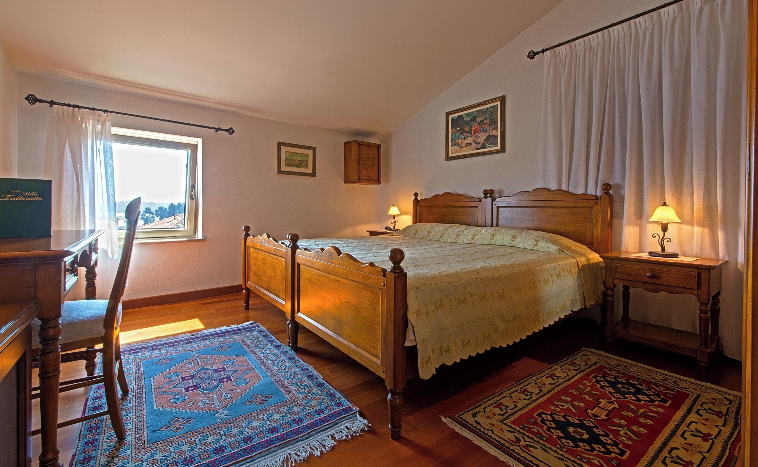 A bedroom with a wooden bed, two bedside tables with lamps, a chair, a window with curtains, and a rug.
