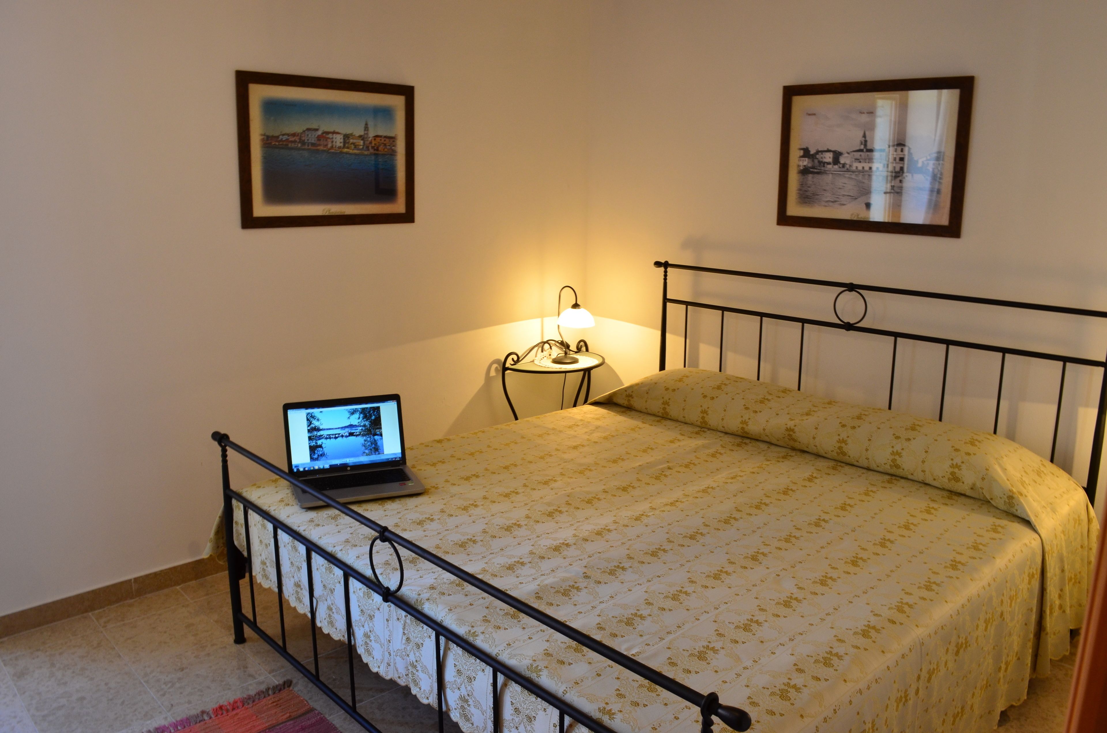 Un dormitorio con una cama, un ordenador portátil al lado, una mesita de noche con una lámpara y dos cuadros en la pared.