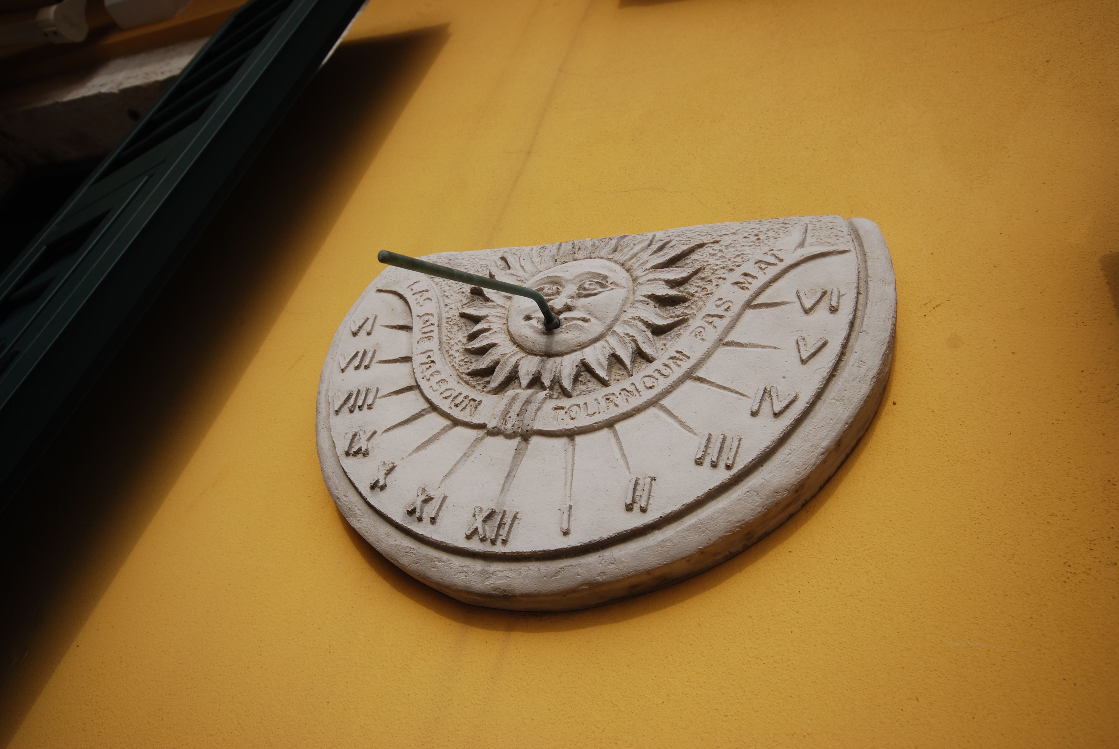 A sundial with Roman numerals and a face on the dial, mounted on a yellow wall.