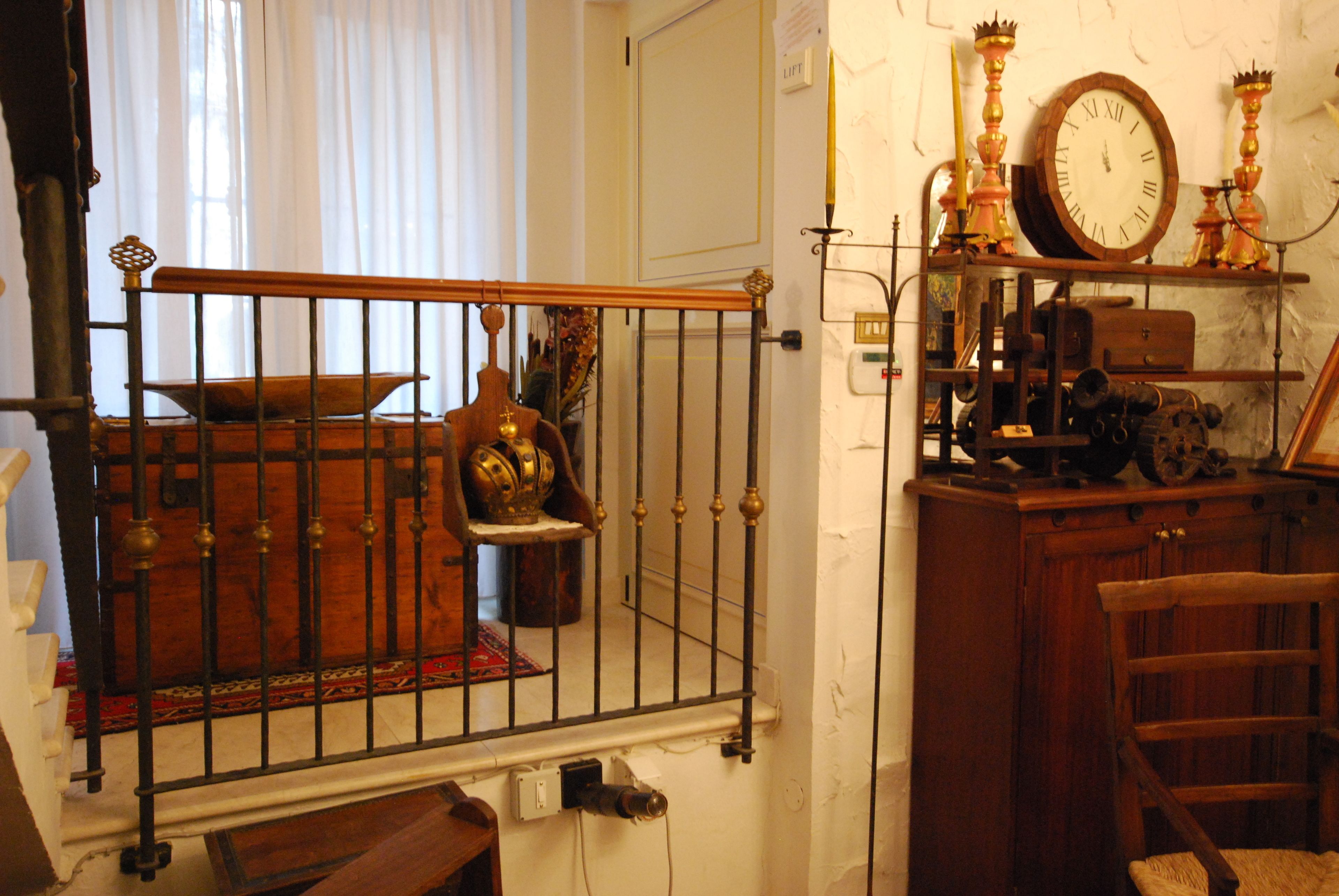 Ein Zimmer mit einem Holzstuhl, einem Metallgeländer, einer Uhr und verschiedenen antiken Gegenständen auf Regalen.
