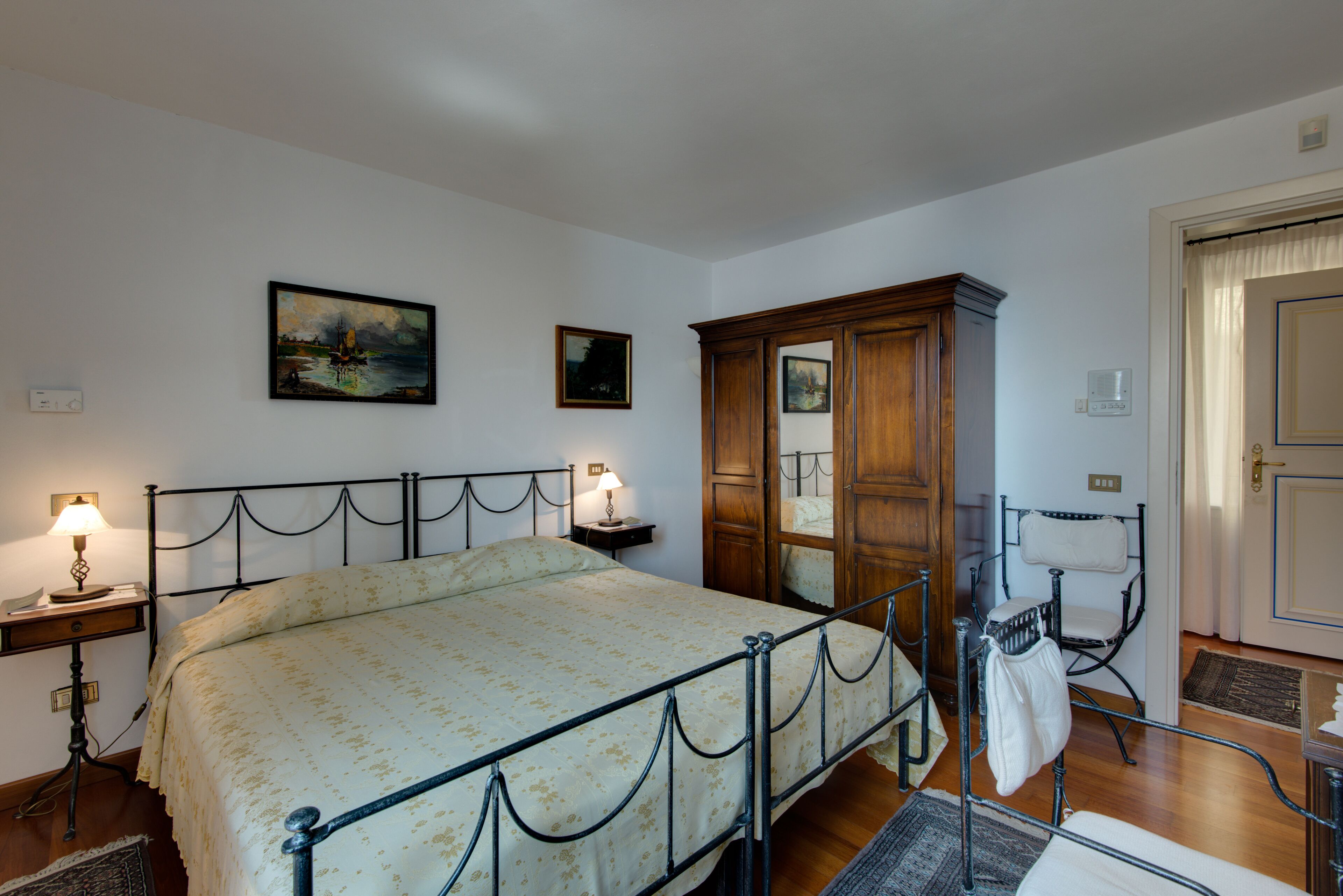 A bedroom with a bed, a nightstand, a lamp, a wooden wardrobe, a chair, and a door.