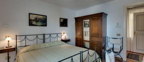A bedroom with a bed, a nightstand, a lamp, a wooden wardrobe, a chair, and a door.