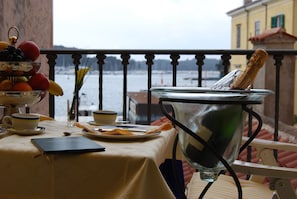 A table set with a fruit bowl, a glass of champagne, and a notebook on a balcony overlooking a body of water.