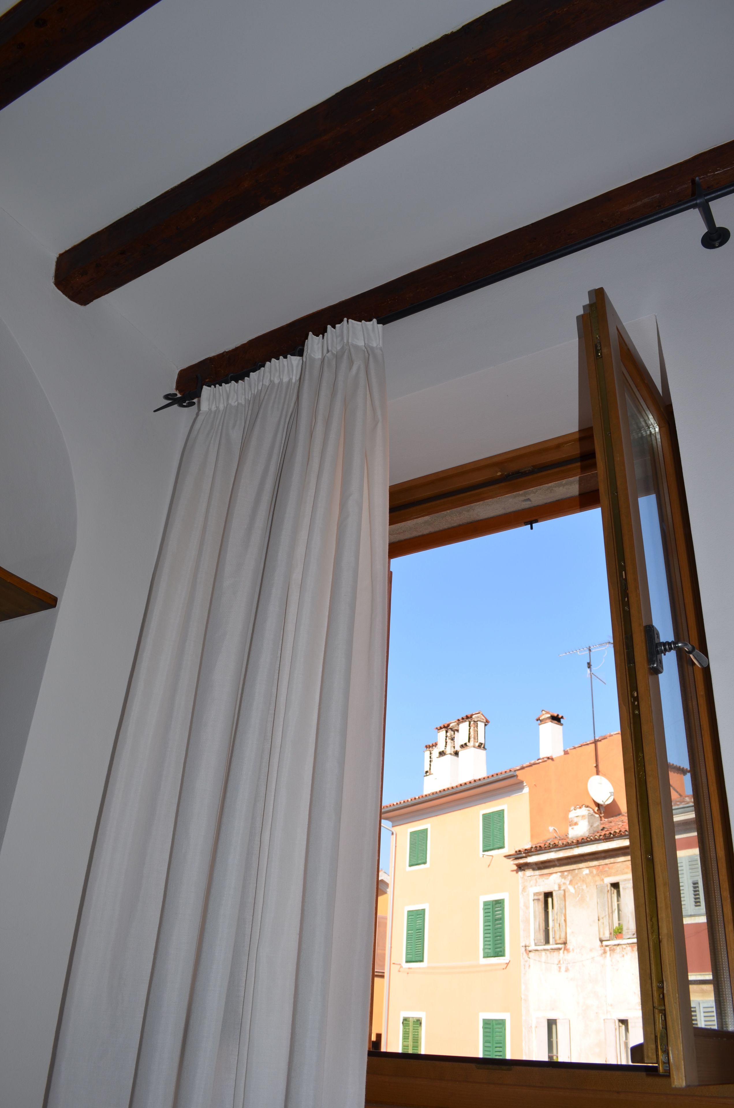 A room with a wooden ceiling beam, a white curtain, and a window with a view of buildings.