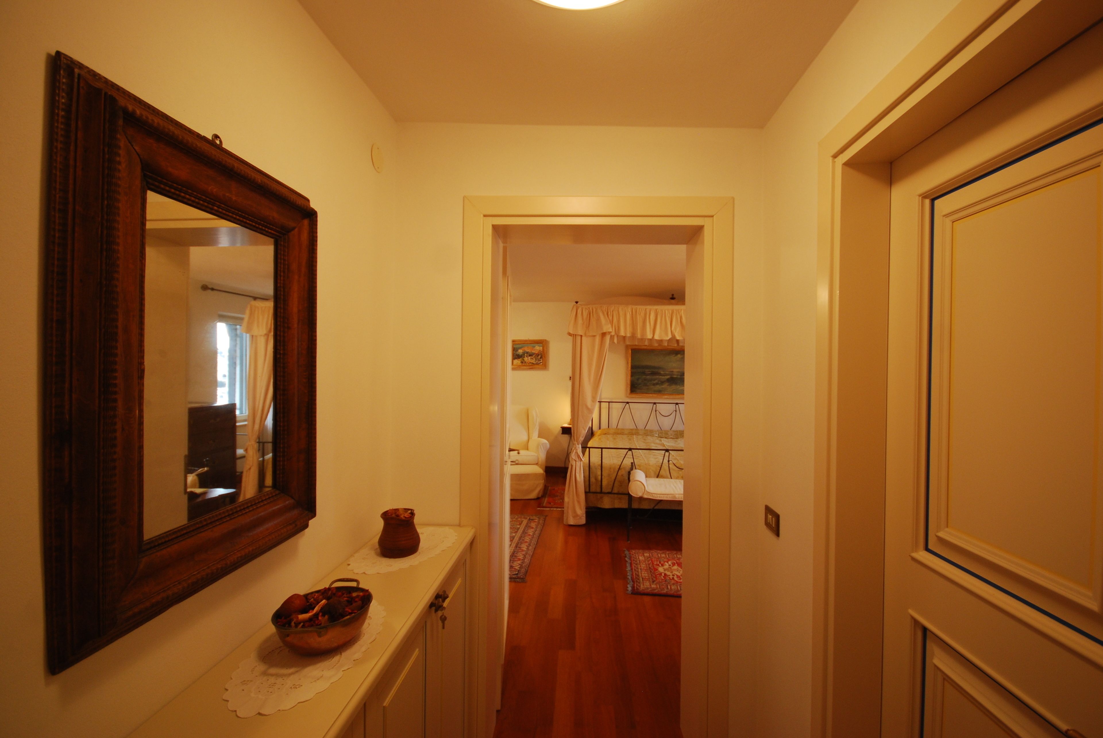 A hallway with a mirror, a small table with a vase, and a door leading to a bedroom with a bed and curtains.