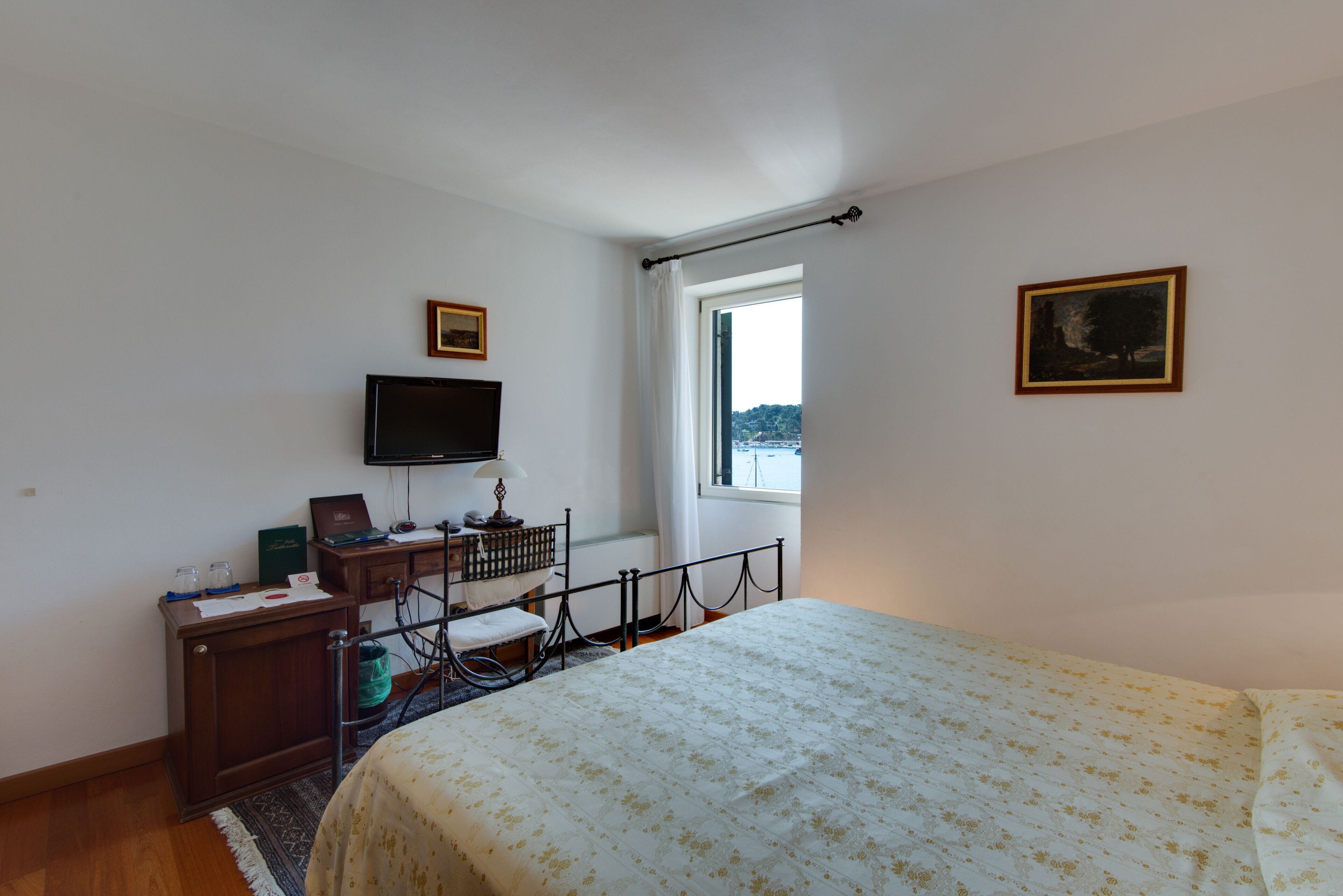A bedroom with a bed, a desk with a computer, a TV, a window with curtains, and two framed pictures on the wall.