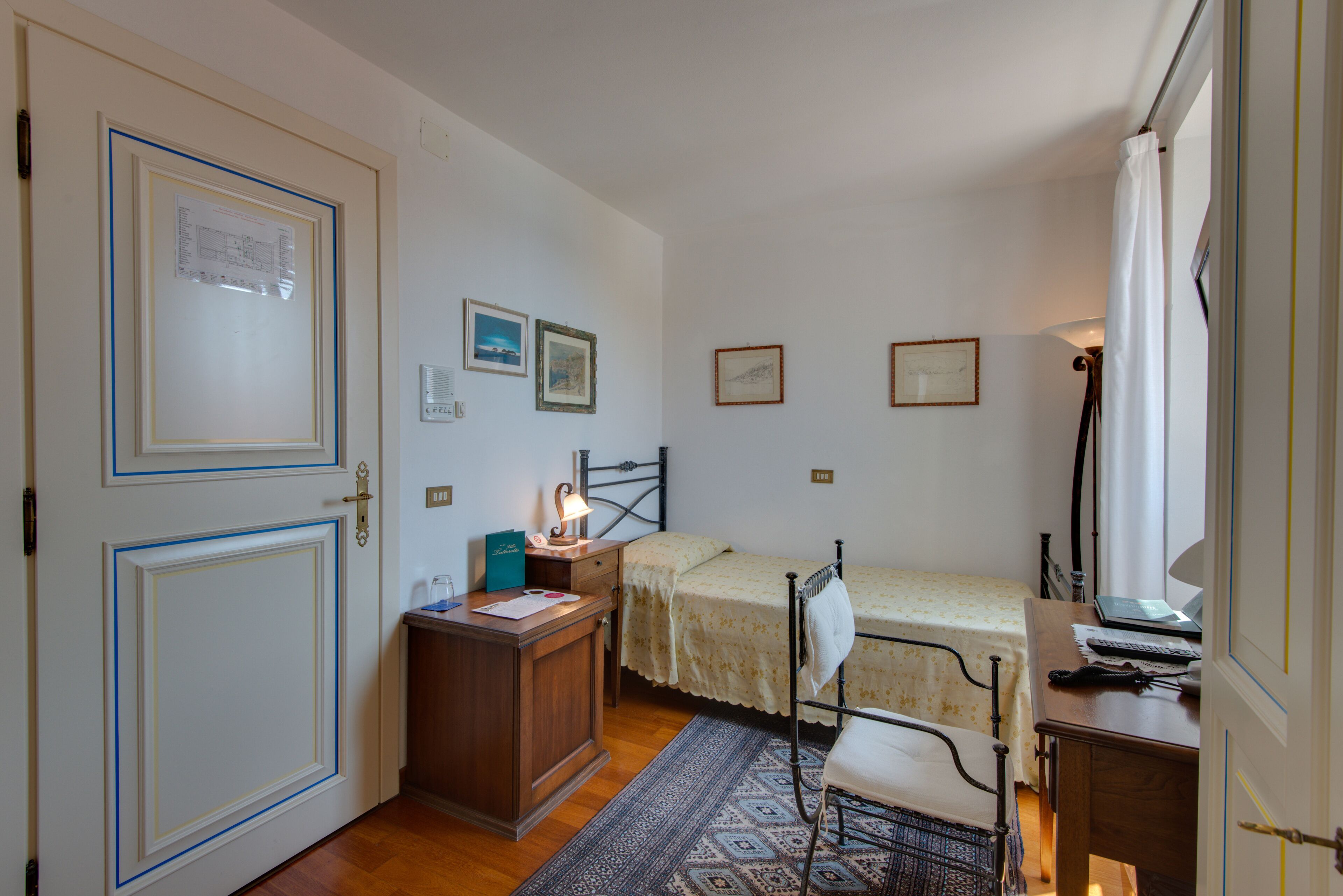 A small, well-lit bedroom with a single bed, a desk, and a chair. There are framed pictures on the wall, a door with a notice, and a window with a curtain.