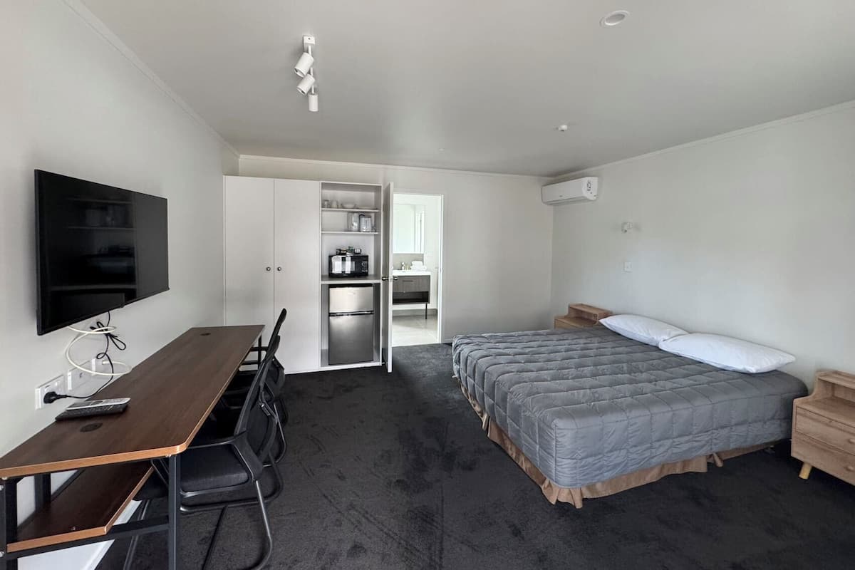 A compact hotel room with a bed, a desk with chairs, a TV, and a kitchenette.