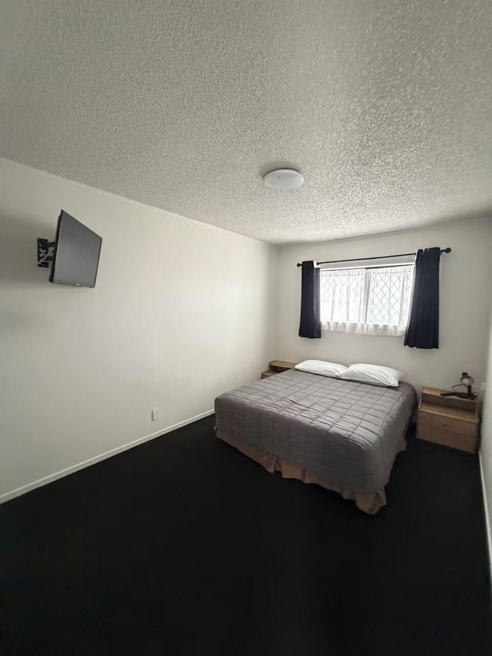 Een slaapkamer met een bed, nachtkastje, een tv aan de muur en een raam met gordijnen.