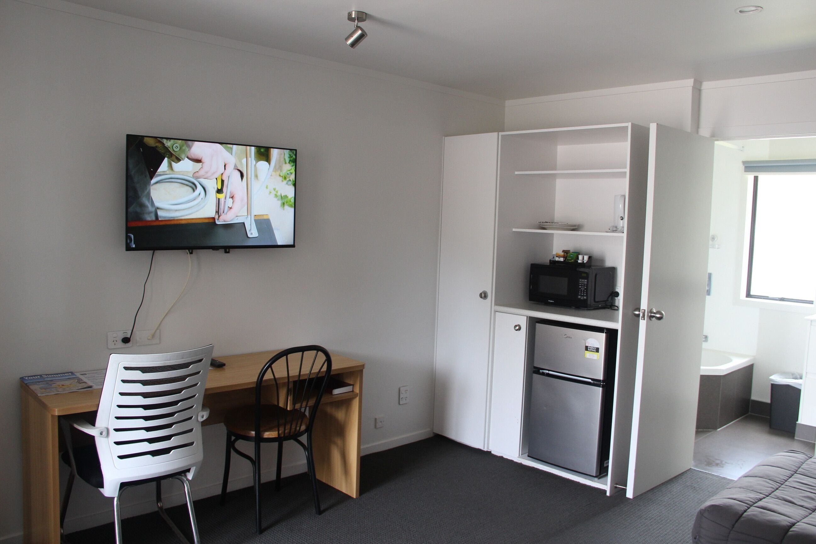 Una habitación compacta con escritorio, silla, televisor, microondas y un refrigerador pequeño.