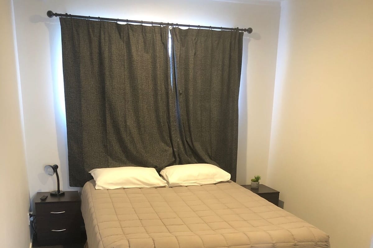 A bedroom with a bed, dark curtains, a bedside table with a lamp, and a small plant.