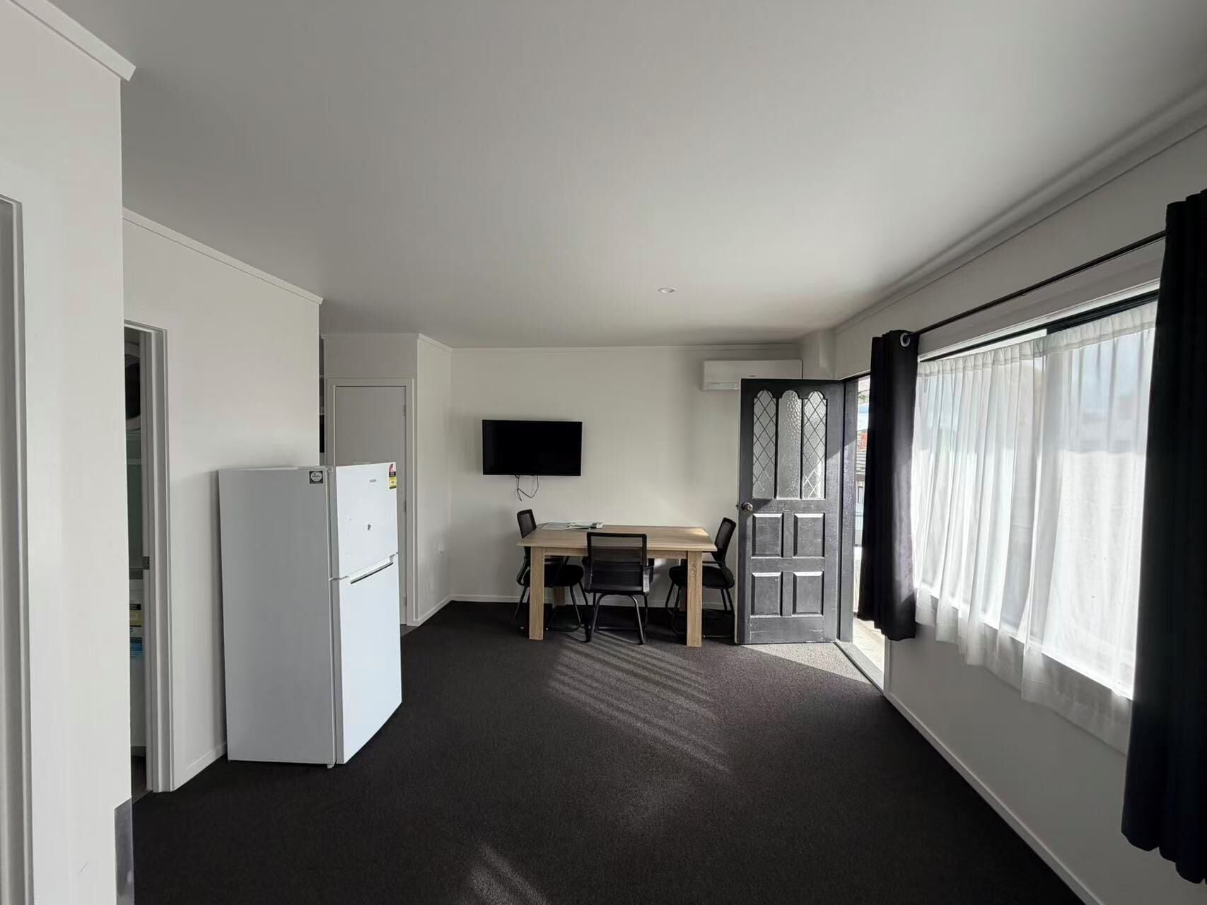 Een kamer met een witte koelkast, een flatscreen televisie aan de muur, een houten eettafel met stoelen en een groot raam met gordijnen.