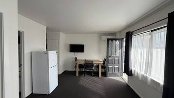 Een kamer met een witte koelkast, een flatscreen televisie aan de muur, een houten eettafel met stoelen en een groot raam met gordijnen.