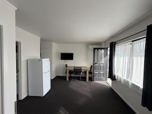 Una habitación con un refrigerador blanco, un televisor de pantalla plana montado en la pared, una mesa de madera con sillas y un ventanal con cortinas.