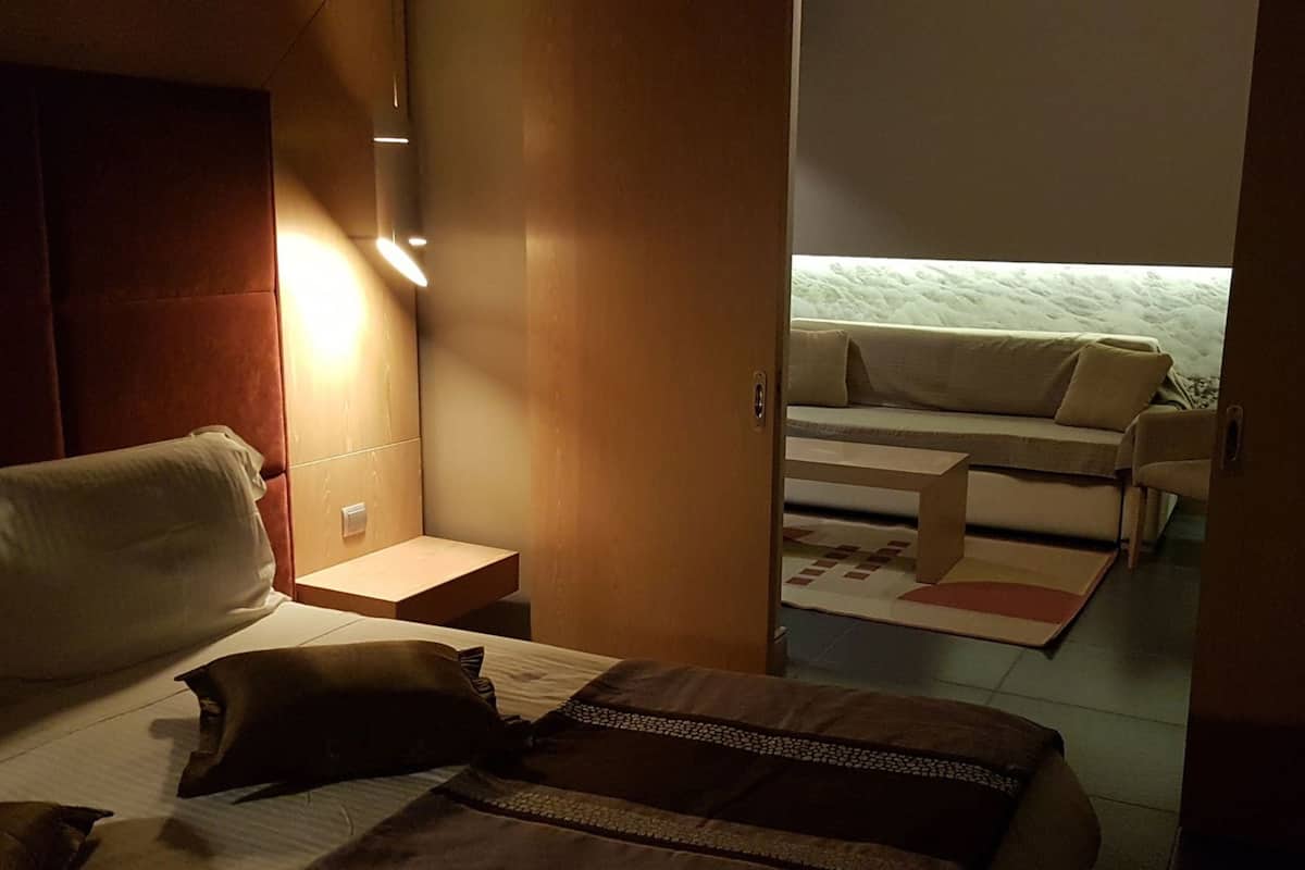A modern hotel room with a bed, bedside table, and a view of a living area with a sofa and chairs.
