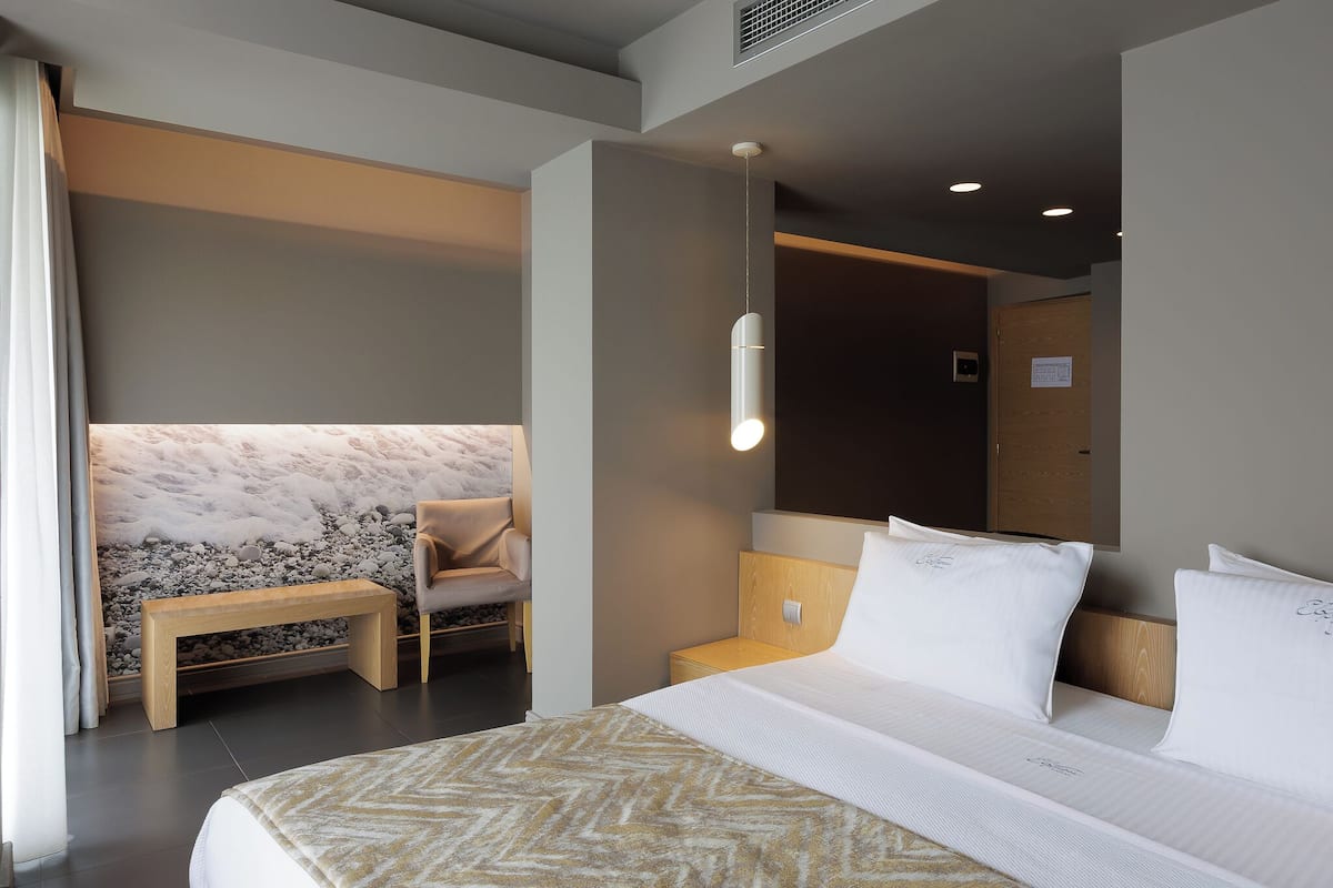 A modern hotel room with a large bed, a bench, a seating area with a wall-mounted light, and a visible door.