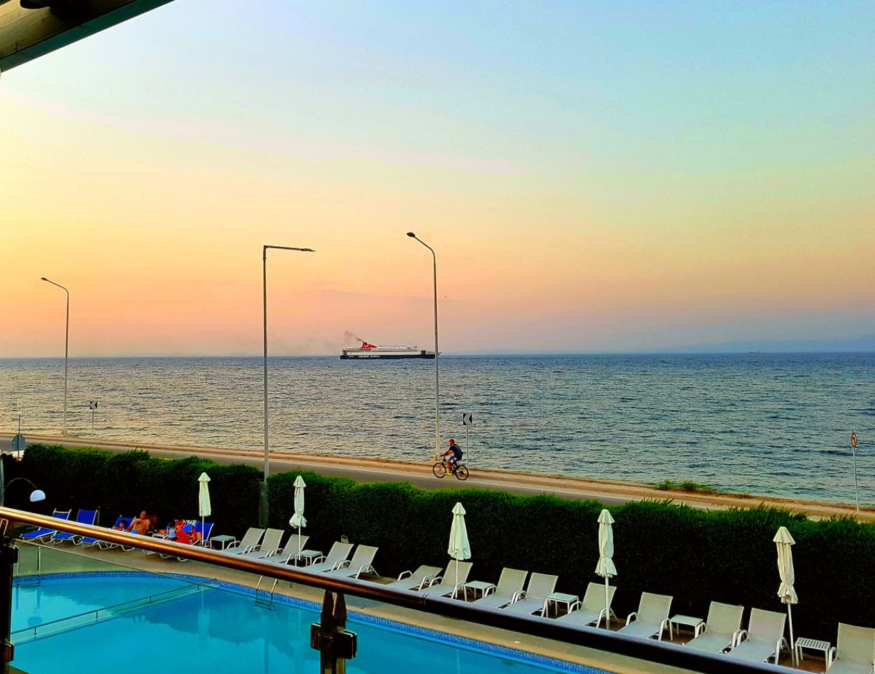 A poolside view with lounge chairs and umbrellas, a road with a cyclist, and a ship in the distance.