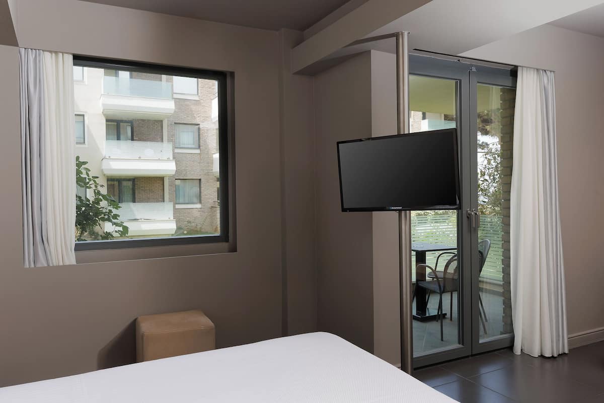 A modern hotel room with a bed, a TV mounted on a sliding glass door, and a view of a building outside.