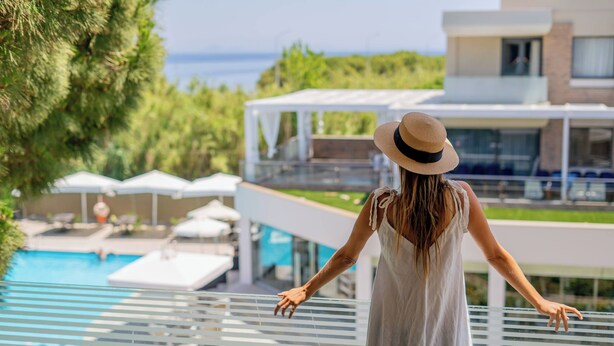 Una persona está de pie en un balcón con vistas a una piscina y un edificio moderno, con una vista despejada del mar.