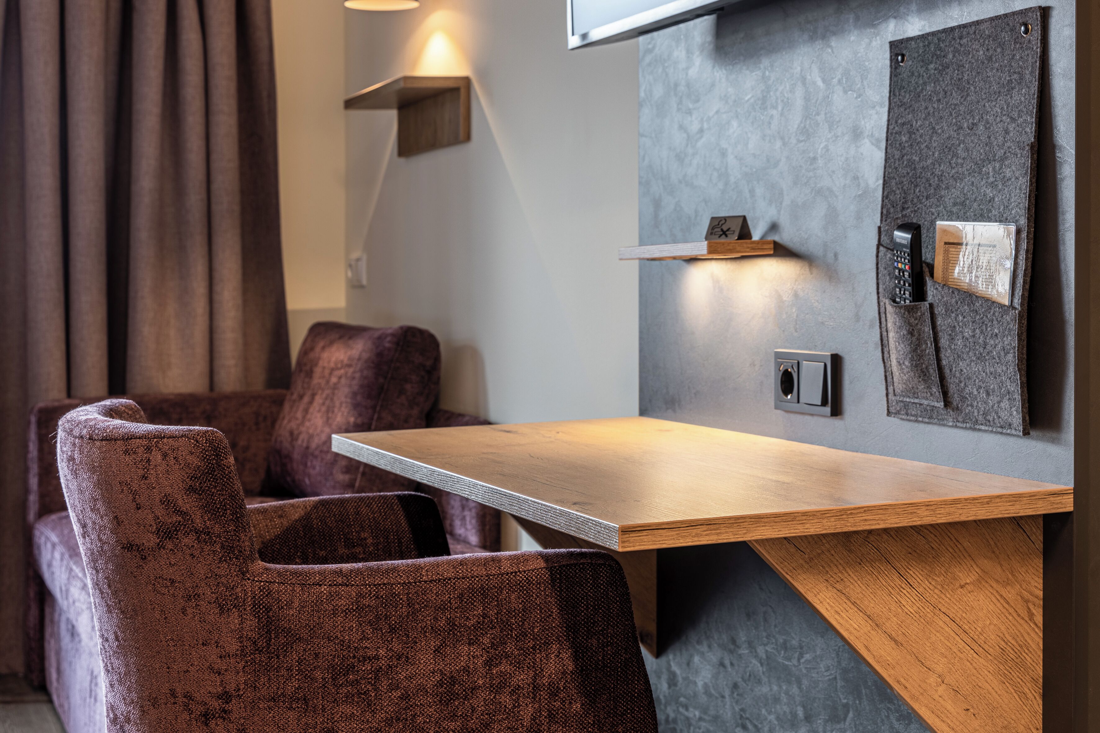 Una habitación de hotel moderna con una mesa de madera, sillas tapizadas de color púrpura, un teléfono en la pared y un pequeño estante con una lámpara.