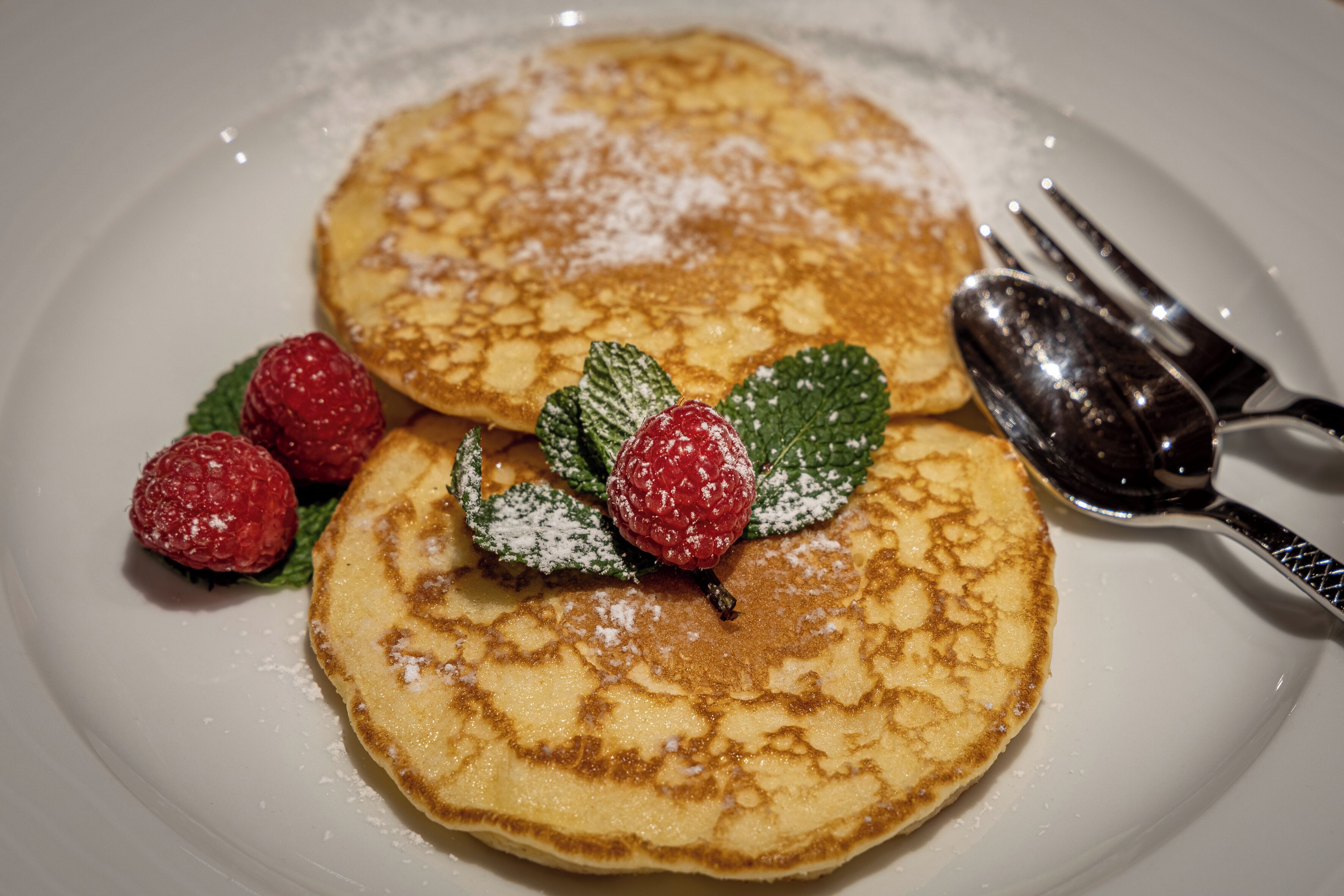 Two pancakes on a white plate with powdered sugar, accompanied by raspberries and mint leaves.