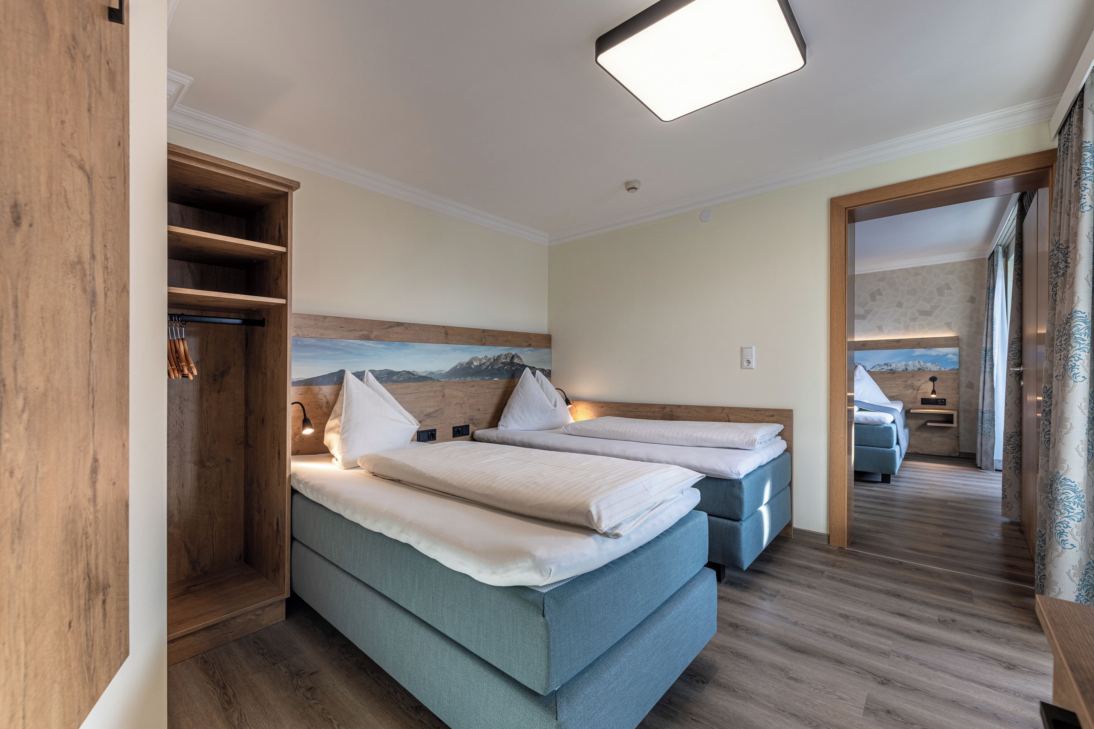 A hotel room with two beds, a wooden wardrobe, a painting on the wall, and a view of the mountains through the window.