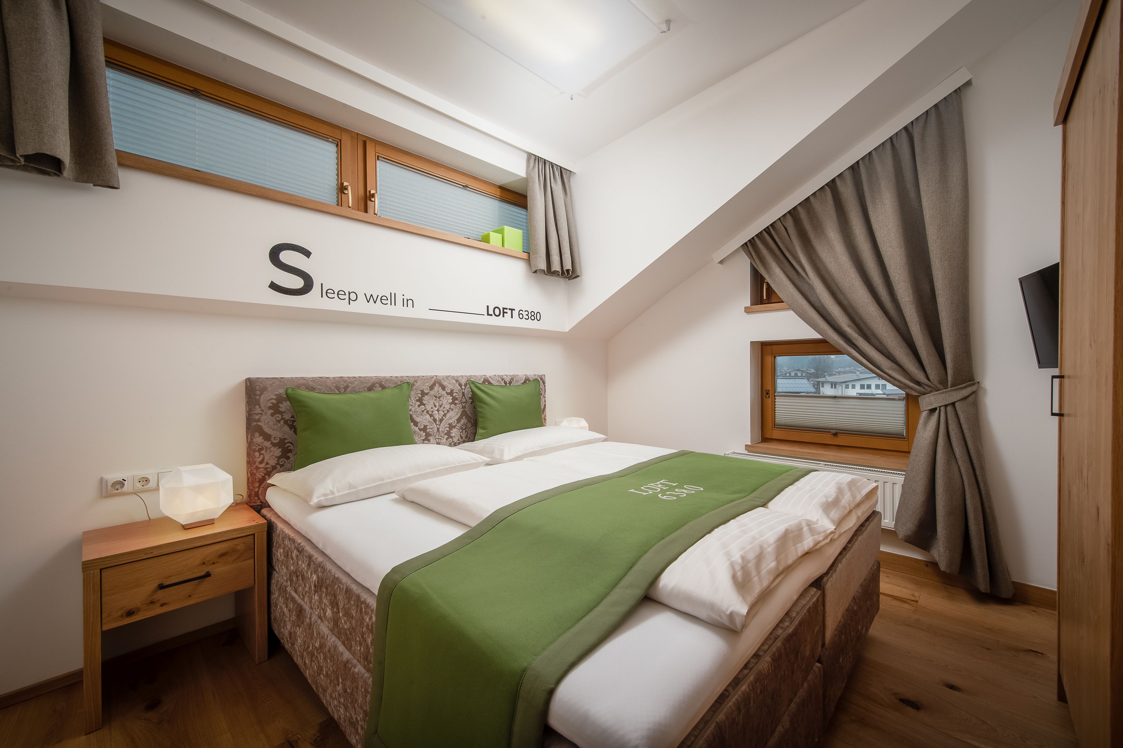 A bedroom with a loft bed, a bedside table, a nightstand, a window with curtains, and a wall-mounted TV.