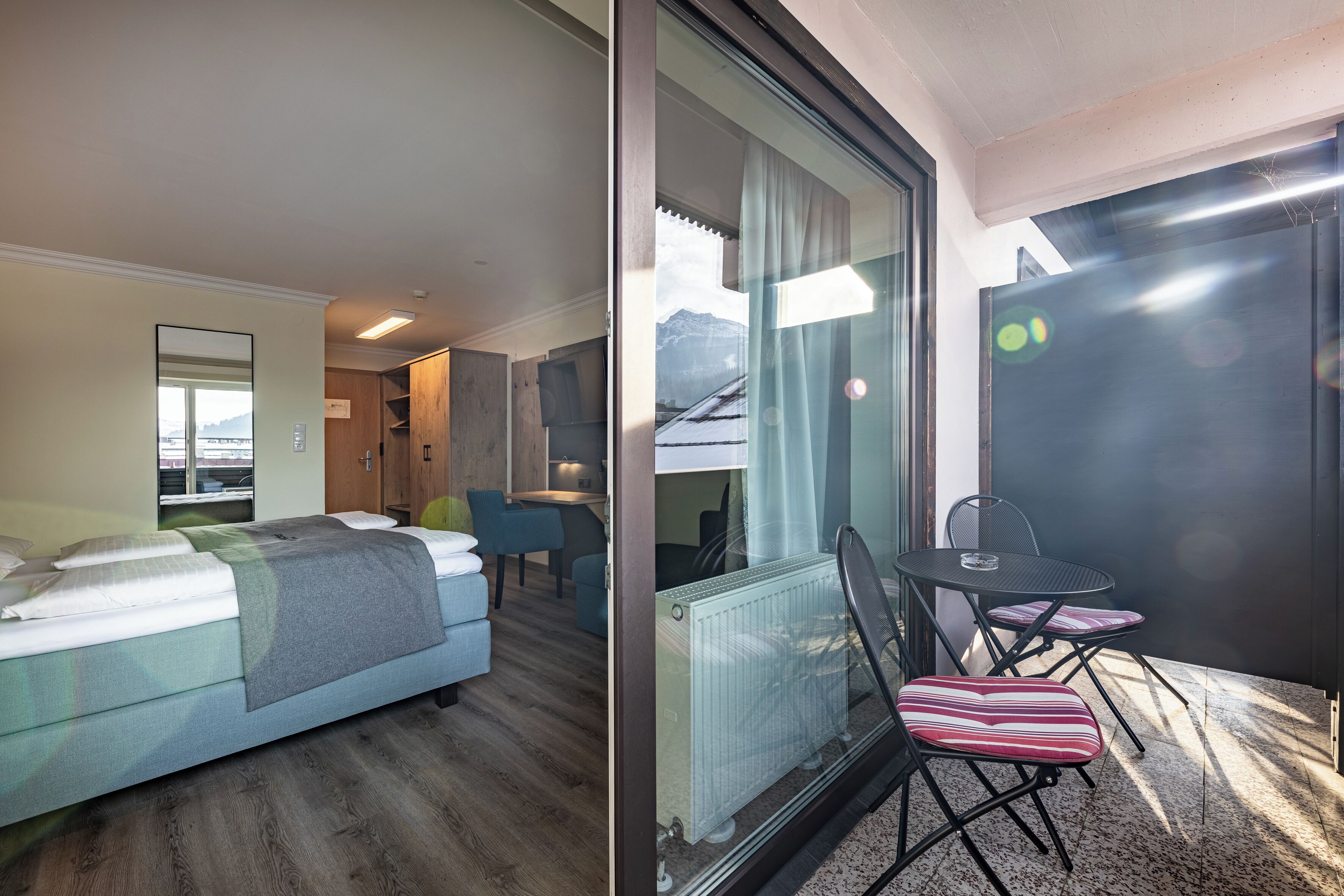 Ein Hotelzimmer mit einem Bett, einem kleinen Tisch mit Stühlen, Bergblick durch ein Fenster und einer Schiebetür zu einem Balkon.