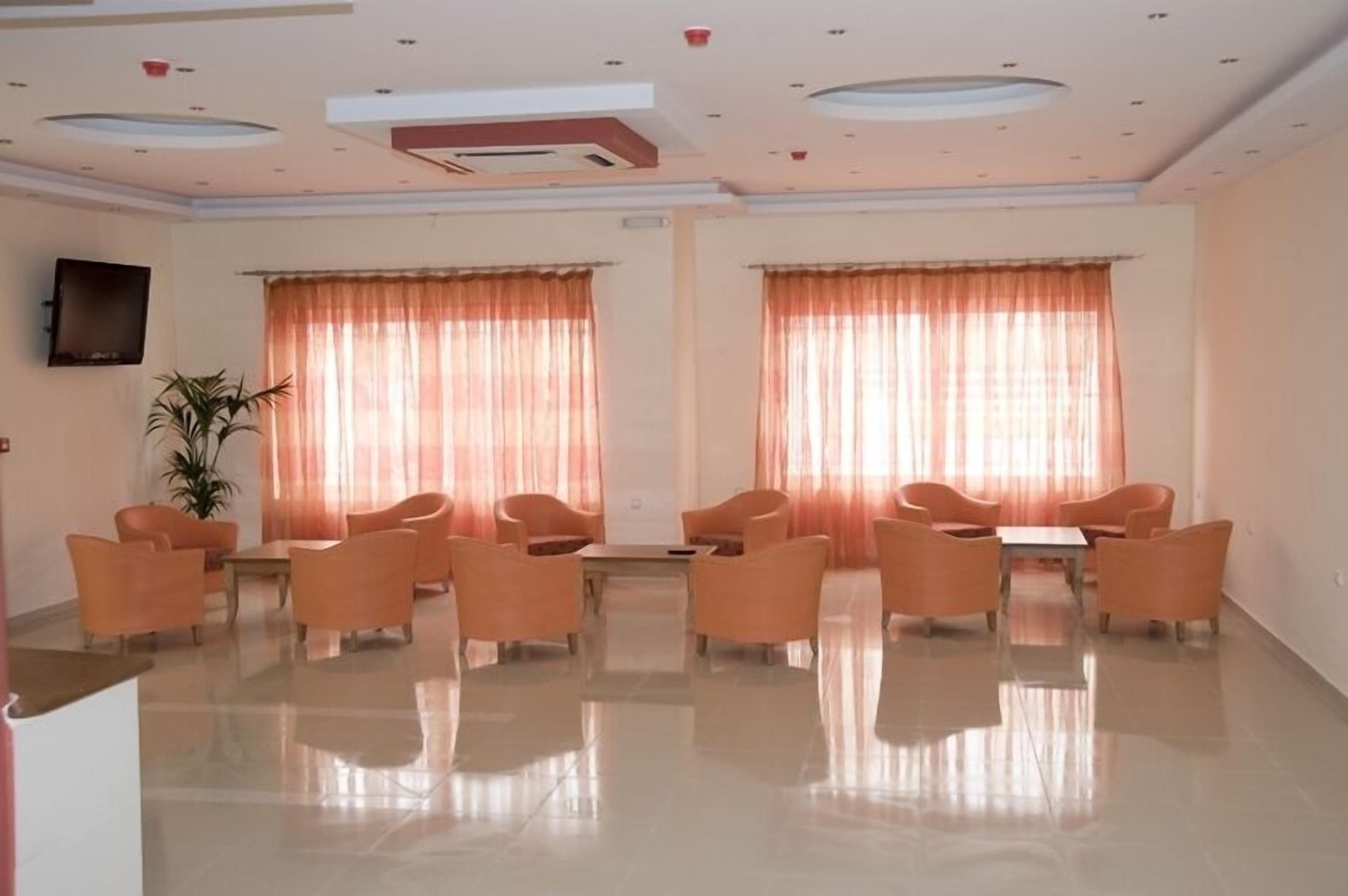 Una habitación amplia con piso pulido, sillas anaranjadas dispuestas alrededor de una mesa central y ventanas grandes con cortinas translúcidas.