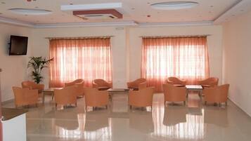 Una habitación amplia con piso pulido, sillas anaranjadas dispuestas alrededor de una mesa central y ventanas grandes con cortinas translúcidas.